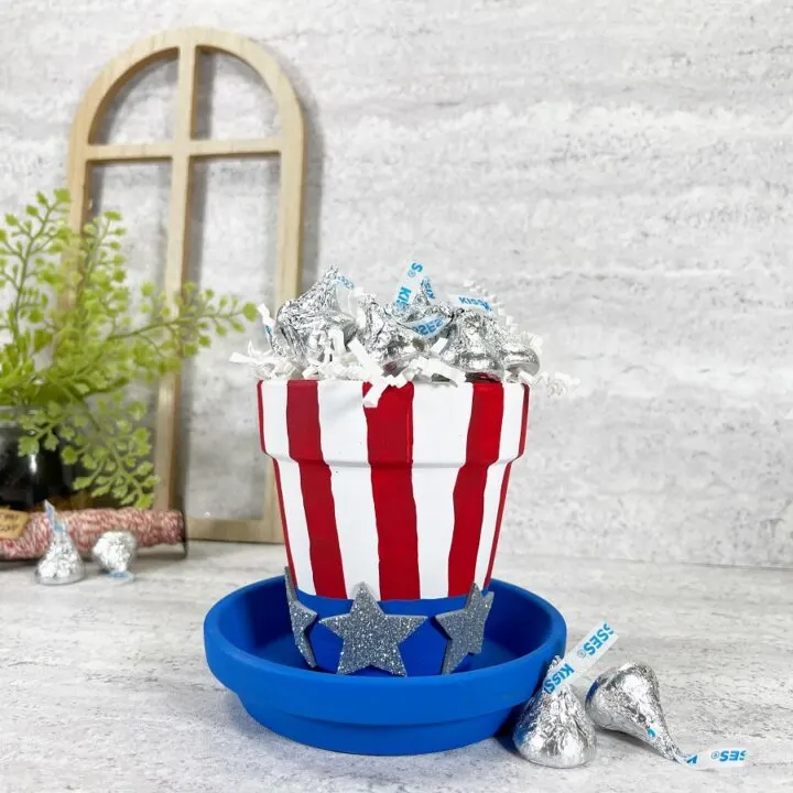 Small clay flower pot painted to look like a patriotic top hat. It is filled with wrapped chocolate kisses. In the background is a small wooden frame and a small green plant.