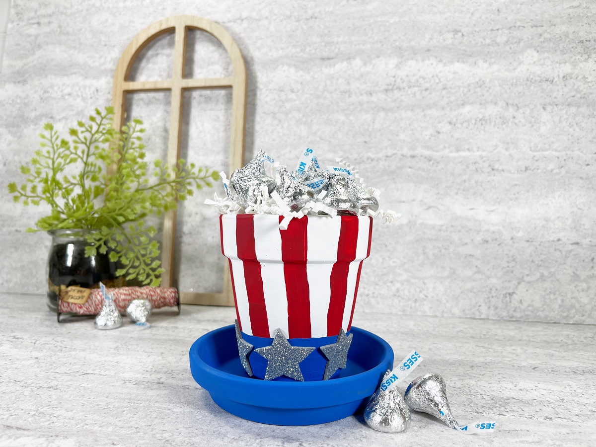 Small clay flower pot painted to look like a patriotic top hat. It is filled with wrapped chocolate kisses. In the background is a small wooden frame and a small green plant.