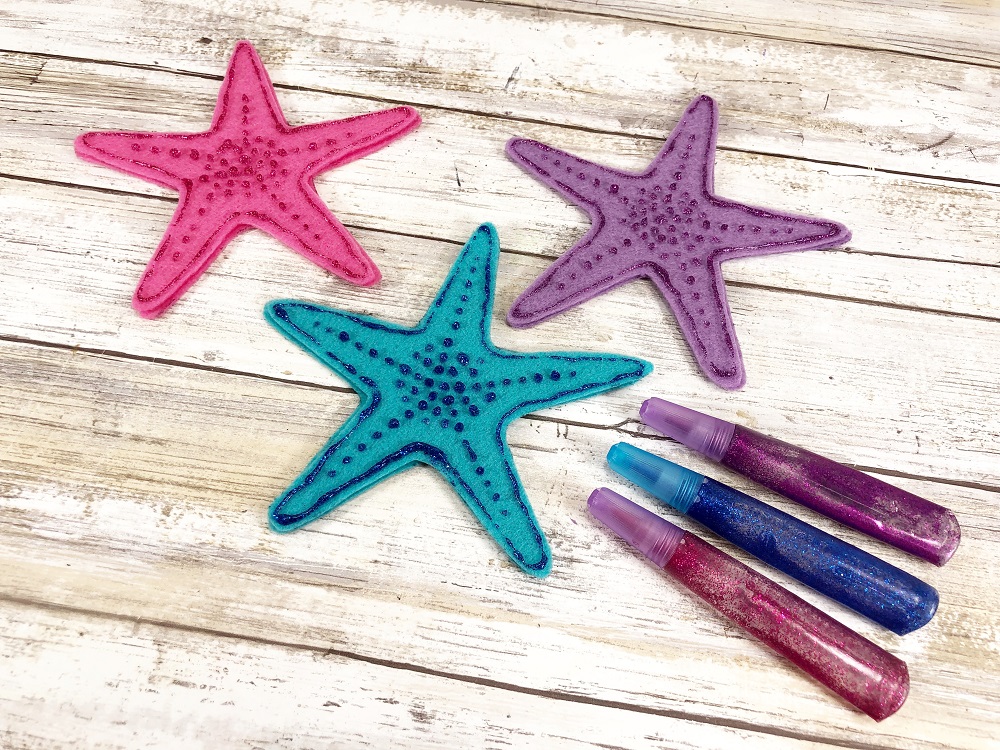 Pink, turquoise, and purple craft felt cut into starfish shapes. Glitter glue pens lay nearby after outlining and dotting details on them.