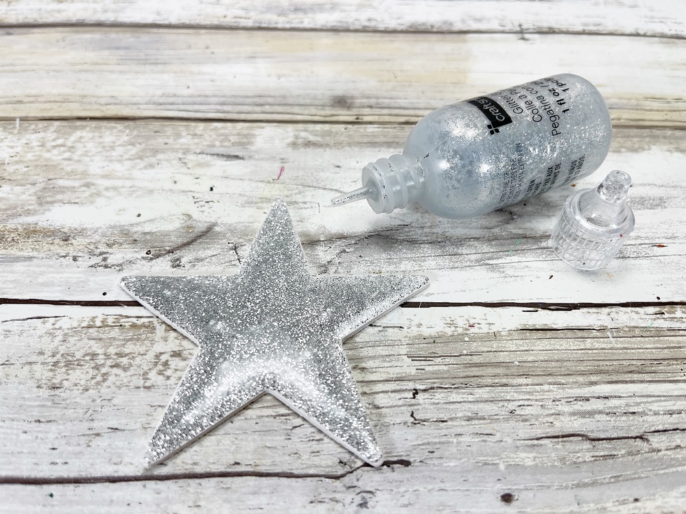 White painted wooden star being covered with a layer of silver glitter glue.