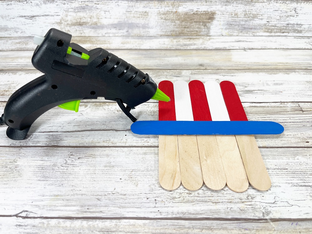 Glue gun next to project after attaching blue painted craft stick across the other ones to create a blue brim on Uncle Sam's hat.