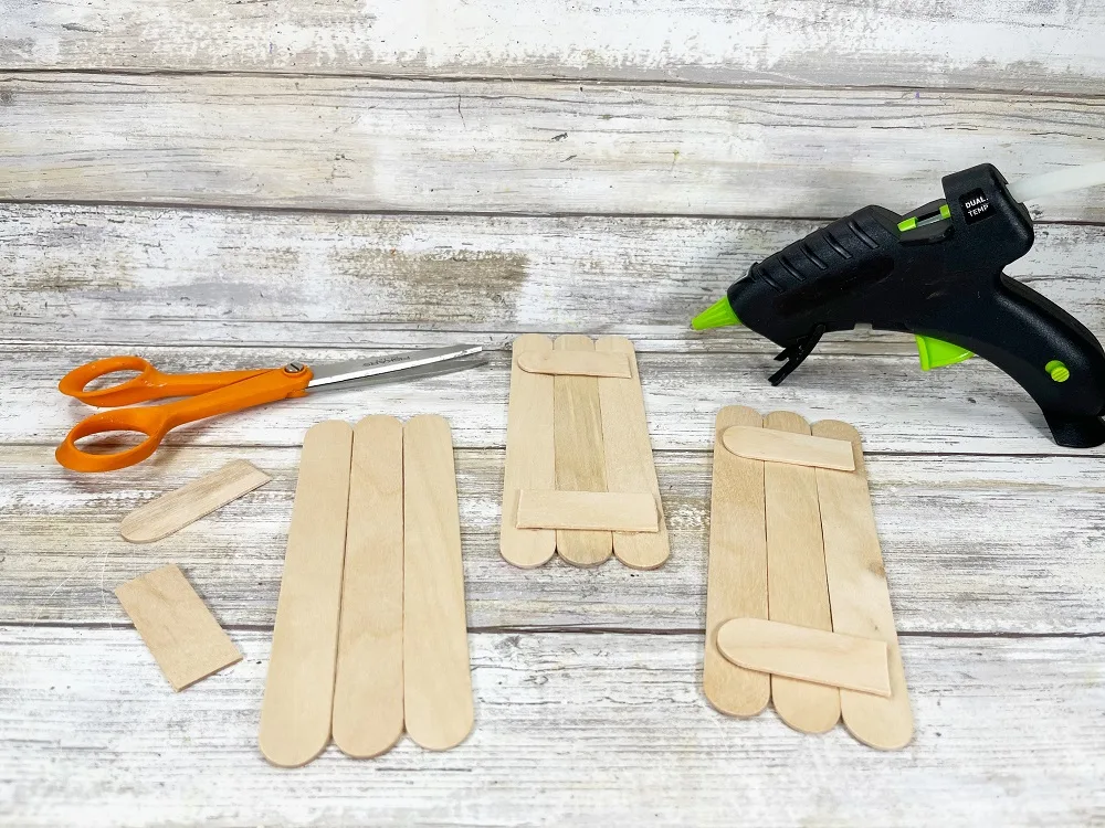 Three groups of jumbo craft sticks in sets of three. Two have pieces of another stick cut and glued across the back with a glue gun.