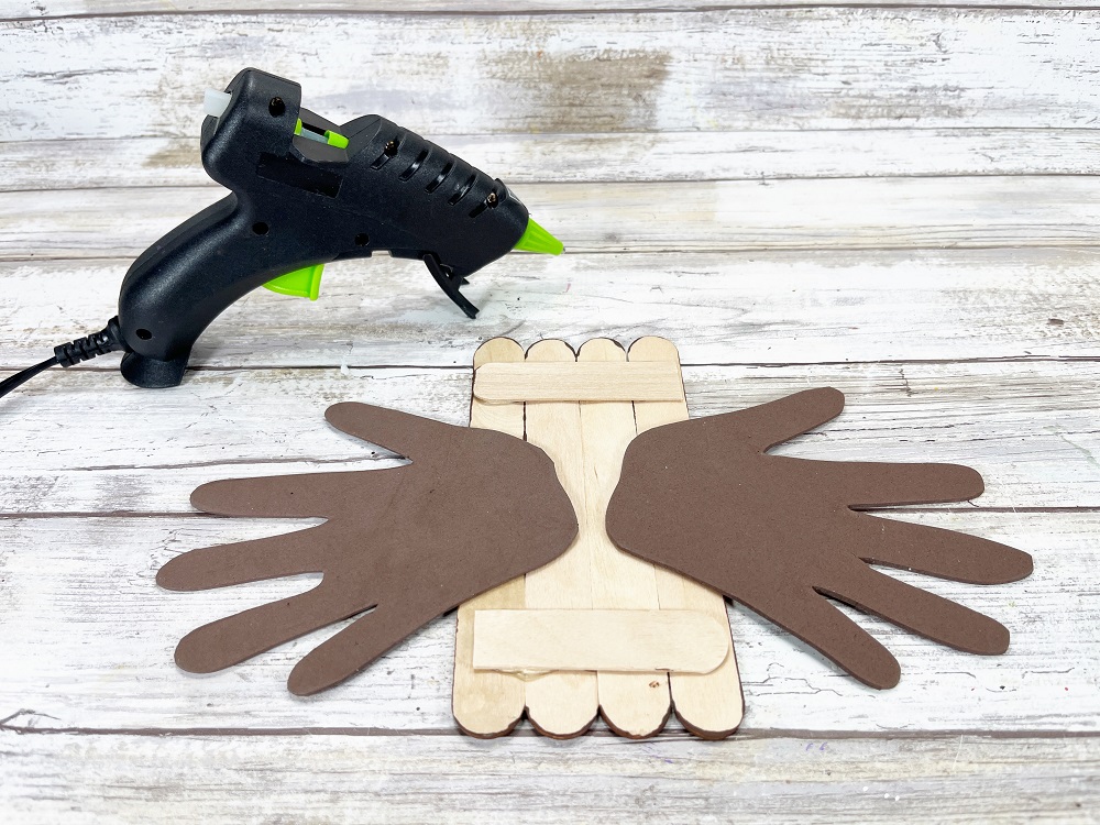 Glue gun sitting near popsicle stick eagle body. It's turned over paint side down. Pair of brown handprint wings glued to the back.