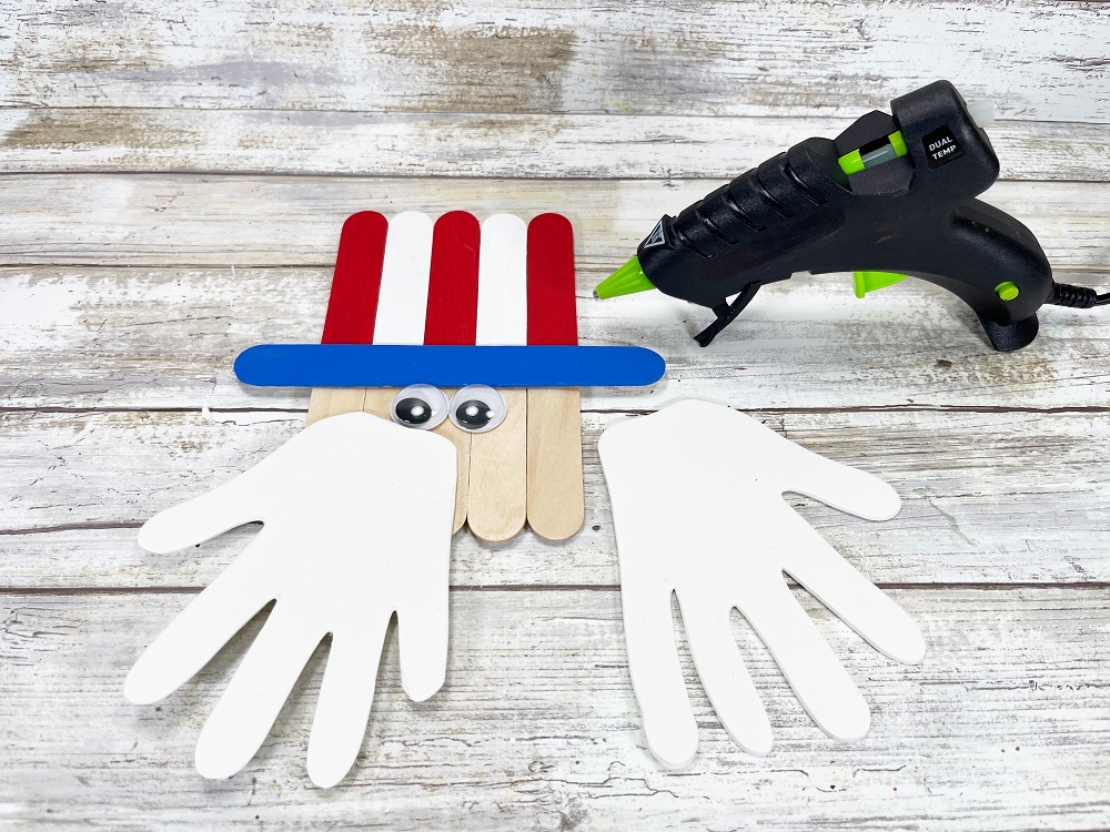 Popsicle stick Uncle Sam with red, white, and blue painted hat. Two googly eyes have been glued onto his face. One white craft foam handprint has been glued on with glue gun. Other handprint off to the side.
