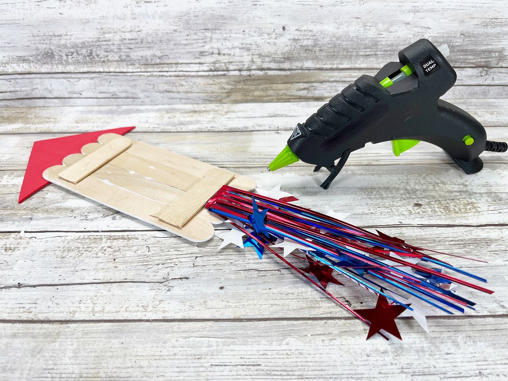 One popsicle stick fireworks rocket flipped over paint side down to show where tinsel flames are being glued in place with hot glue gun.