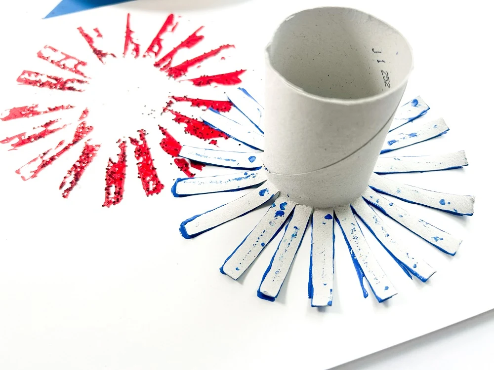 Close view of TP roll stamping blue fireworks onto white paper close to red burst of fireworks.