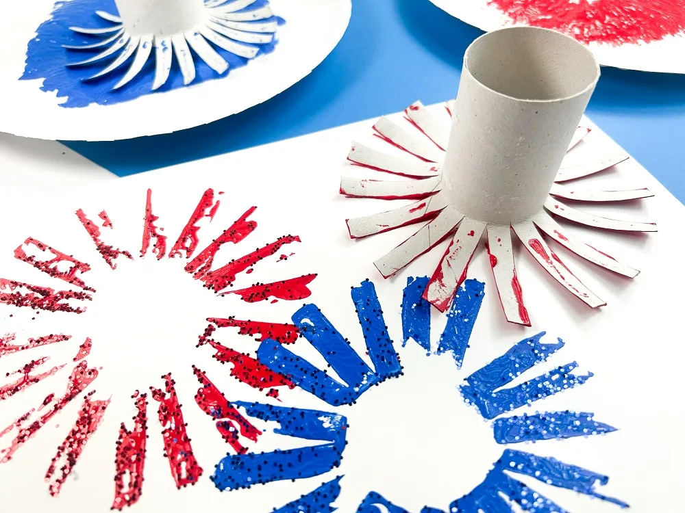 Red and blue fireworks painted on white paper and have glitter sprinkled on them. Toilet paper roll covered in red paint pressed against paper to make another one.