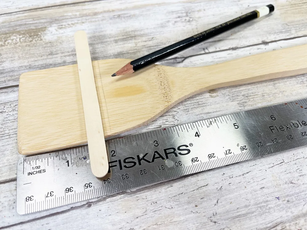 Flat wooden spatula laying horizontally next to a ruler to measure about two inches from the top and mark a line using the craft stick.