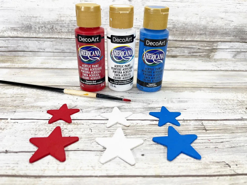 Three pairs of wooden stars are painted red, white, and blue. Paint brush and bottles of paint are behind where the stars are lined up.
