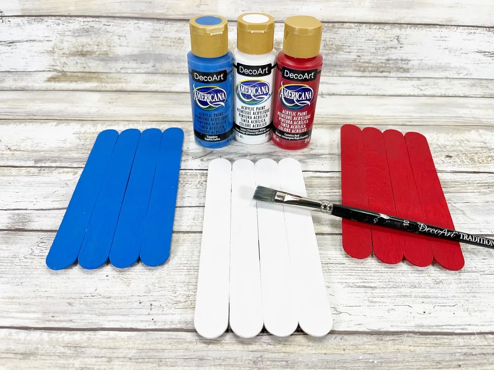 Three different sets of craft sticks have been painted. Left set is blue, middle set is white, and right set is red. Paint bottles and paintbrush are by them.