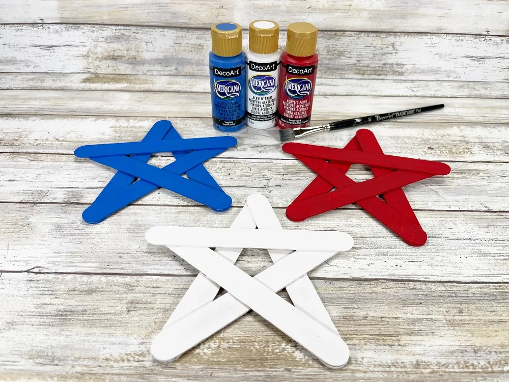Bottles of blue, white, and red craft paint and a paint brush sitting behind where three painted stars made with craft sticks are laid out. One star is painted blue, one white, and the other red.