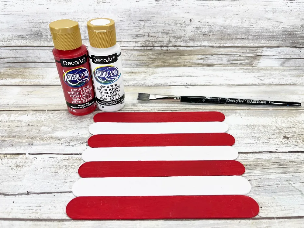 Bottles of red and white paint and paintbrush near jumbo craft sticks. Sticks have been painted and are laid out alternating in red and white like stripes.