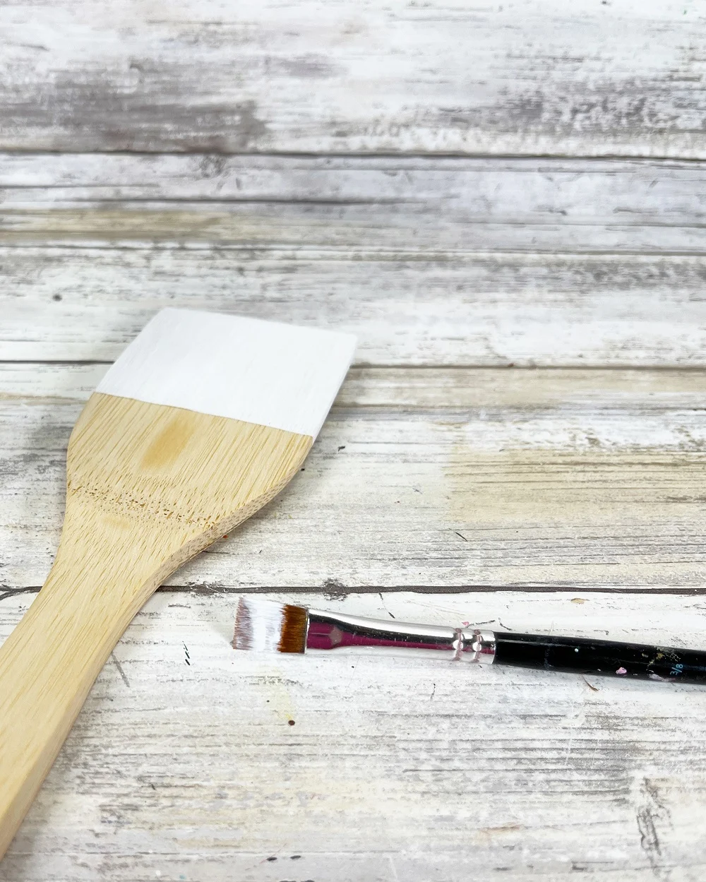 Top part of bamboo spatula partially painted white. Paintbrush with white paint on it laying nearby.