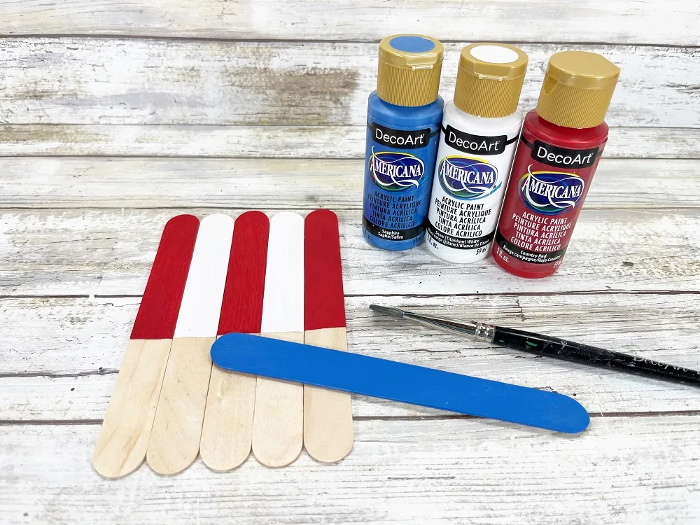Five jumbo craft sticks laying next to each other vertically. Top half painted in alternating red and white. One full blue painted craft stick lays over them. Paint bottles and paintbrush next to craft sticks.