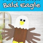 Black and white text on light blue background at top of image says Popsicle Stick Bald Eagle. Underneath is a close view of a hand holding up a finished craft project made with popsicle sticks and craft foam pieces.