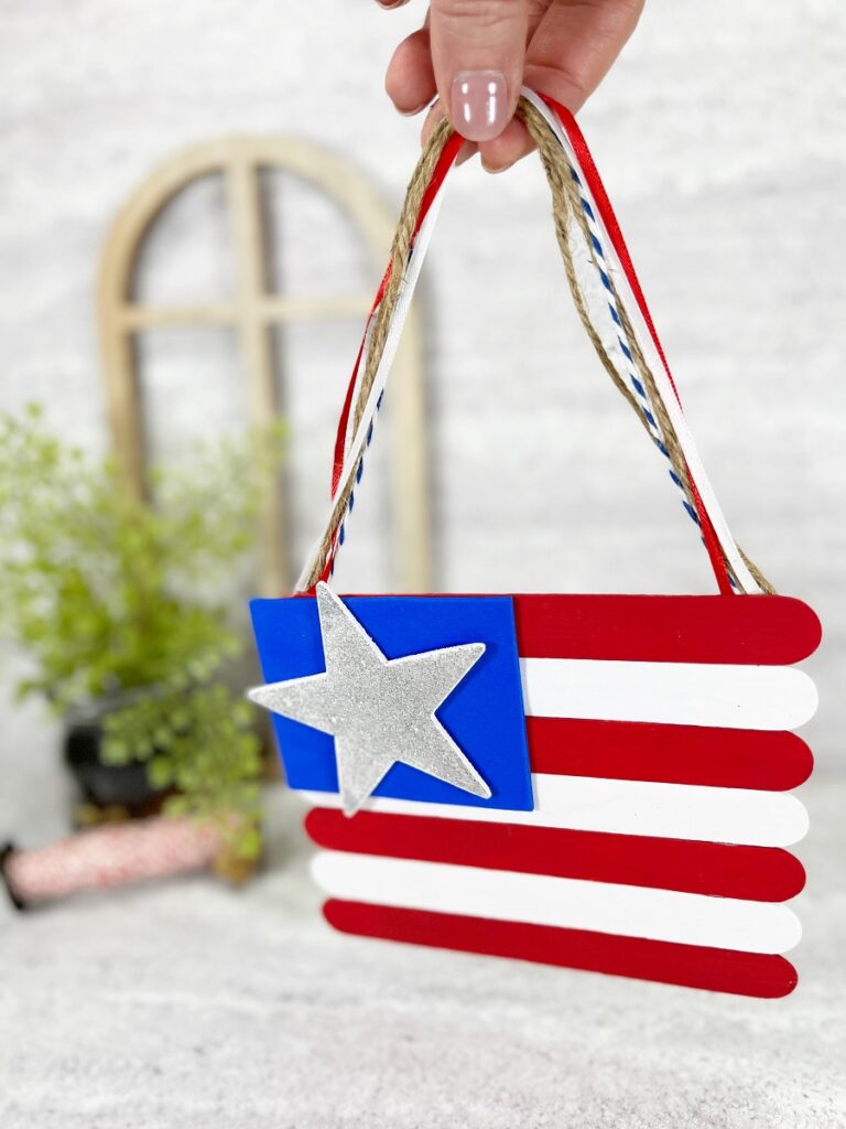 Hand holding a small flag made using craft sticks and made into a hanging decoration using twine and ribbon.