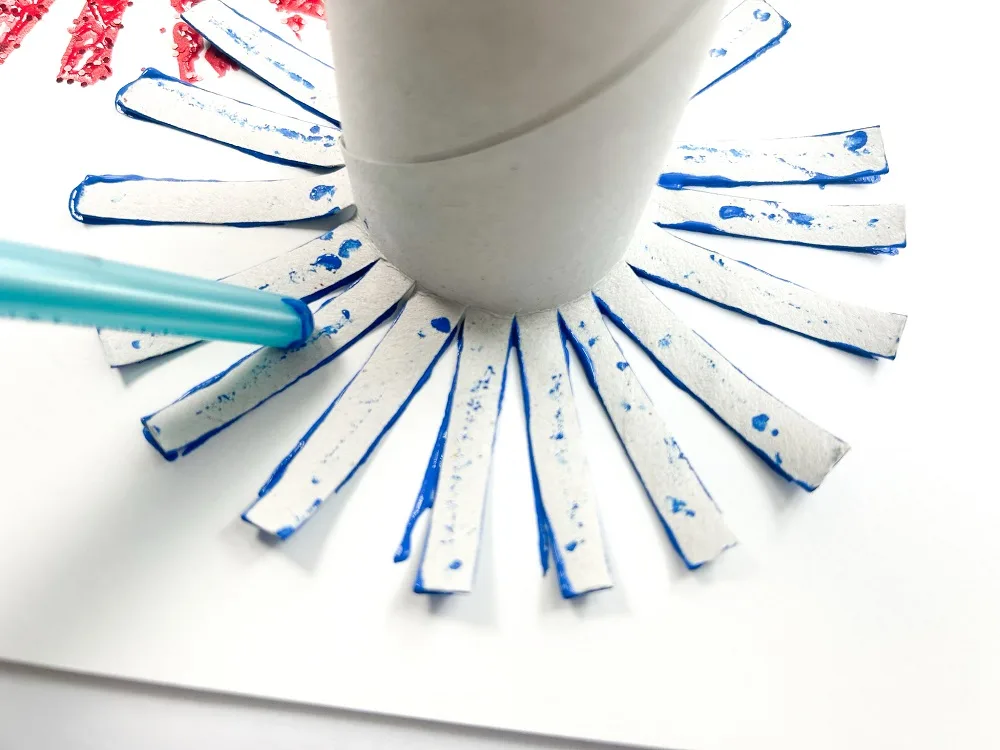 Cut toilet paper roll covered in blue paint being stamped down onto white paper. End of blue paintbrush is pressing the strips down against the paper.