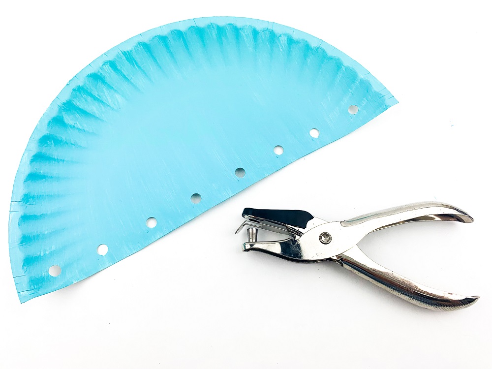 Half of a large paper plate painted blue with holes along the flat side. Silver single hole puncher laying next to plate.