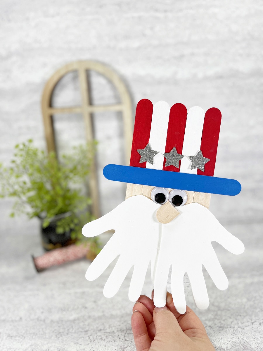 Hand holding up finished craft stick Uncle Sam in right front of picture. Small green plant is blurred in the background.