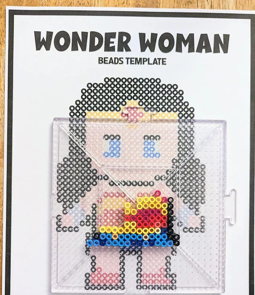 Overhead view of Wonder Woman inspired bead pattern printed out on paper. Large clear pegboard laying over the design with some of the beads filled in the middle.