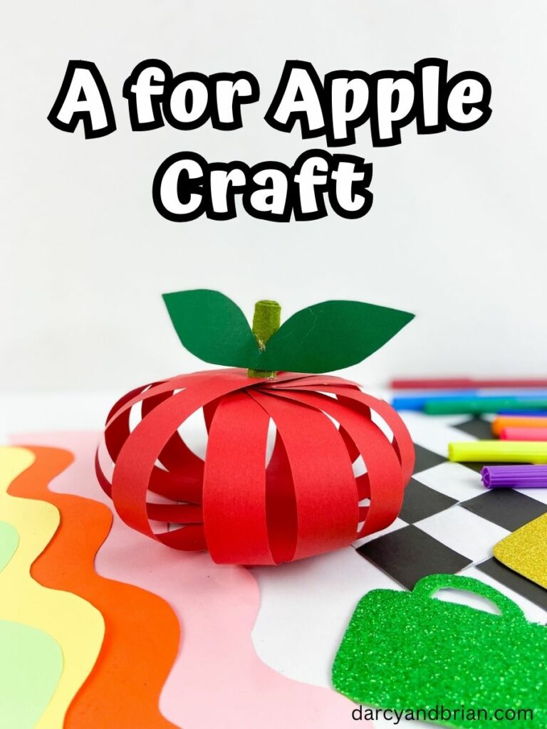 White text with thick black outline says A for Apple Craft above apple made out of paper strips sitting on top of assorted supplies and colored paper.