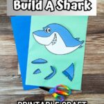 A color version of printable shark craft printed out. Pieces are cut out and arranged on light green and dark blue construction papers. A child's pair of rainbow handled scissors lays across the bottom. Top has a text box that says Build a Shark.