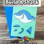 A color version of printable shark craft printed out. Pieces are cut out and arranged on light green and dark blue construction papers. A child's pair of rainbow handled scissors lays across the bottom. Top has a text box that says Build a Shark.