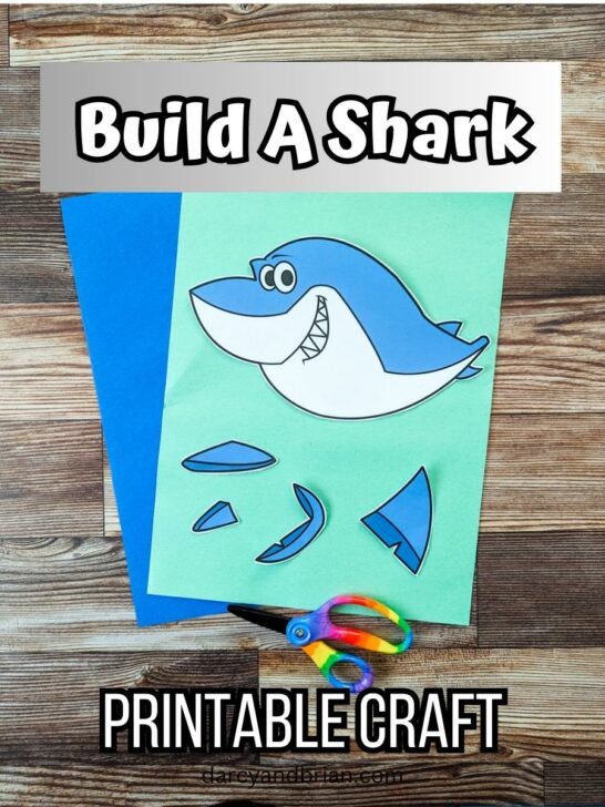 A color version of printable shark craft printed out. Pieces are cut out and arranged on light green and dark blue construction papers. A child's pair of rainbow handled scissors lays across the bottom. Top has a text box that says Build a Shark.