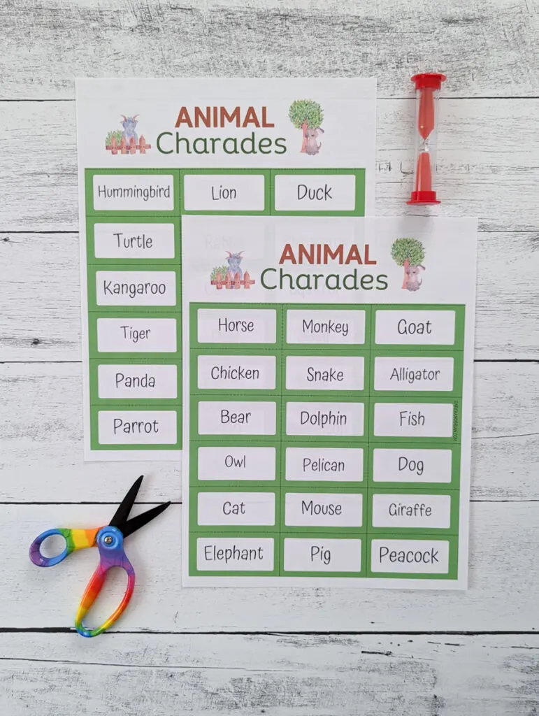 Two pages of animal charades word cards printed out and overlapping each other on a white wood background. A pair of scissors is in the lower left. A red sand timer is in the upper right.