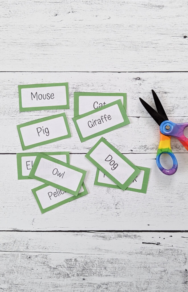 Several charades word cards with different animals typed on them are cut apart and laying on a white wood background next to scissors.