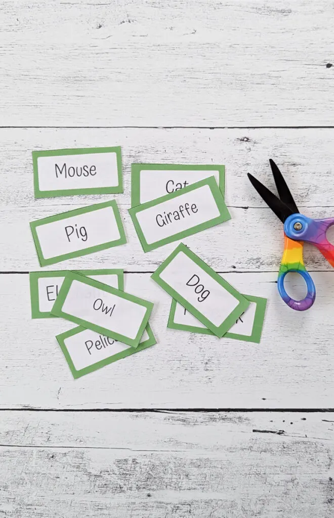 Several charades word cards with different animals typed on them are cut apart and laying on a white wood background next to scissors.