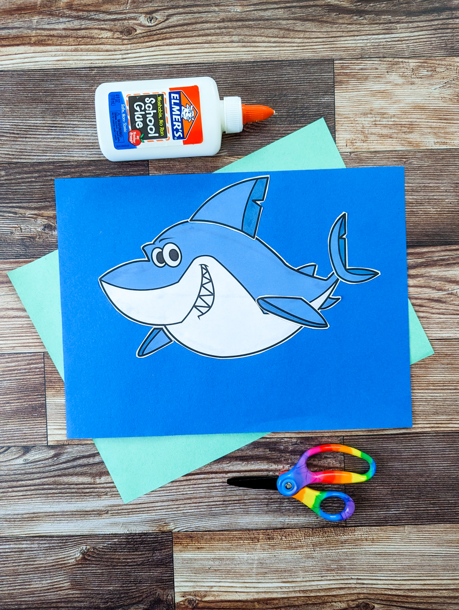 Great White Shark printable cut out and glued into place on blue construction paper. Piece of light green construction paper lays underneath it. Bottle of glue lays along the top of the craft and kid scissors lay along the bottom.