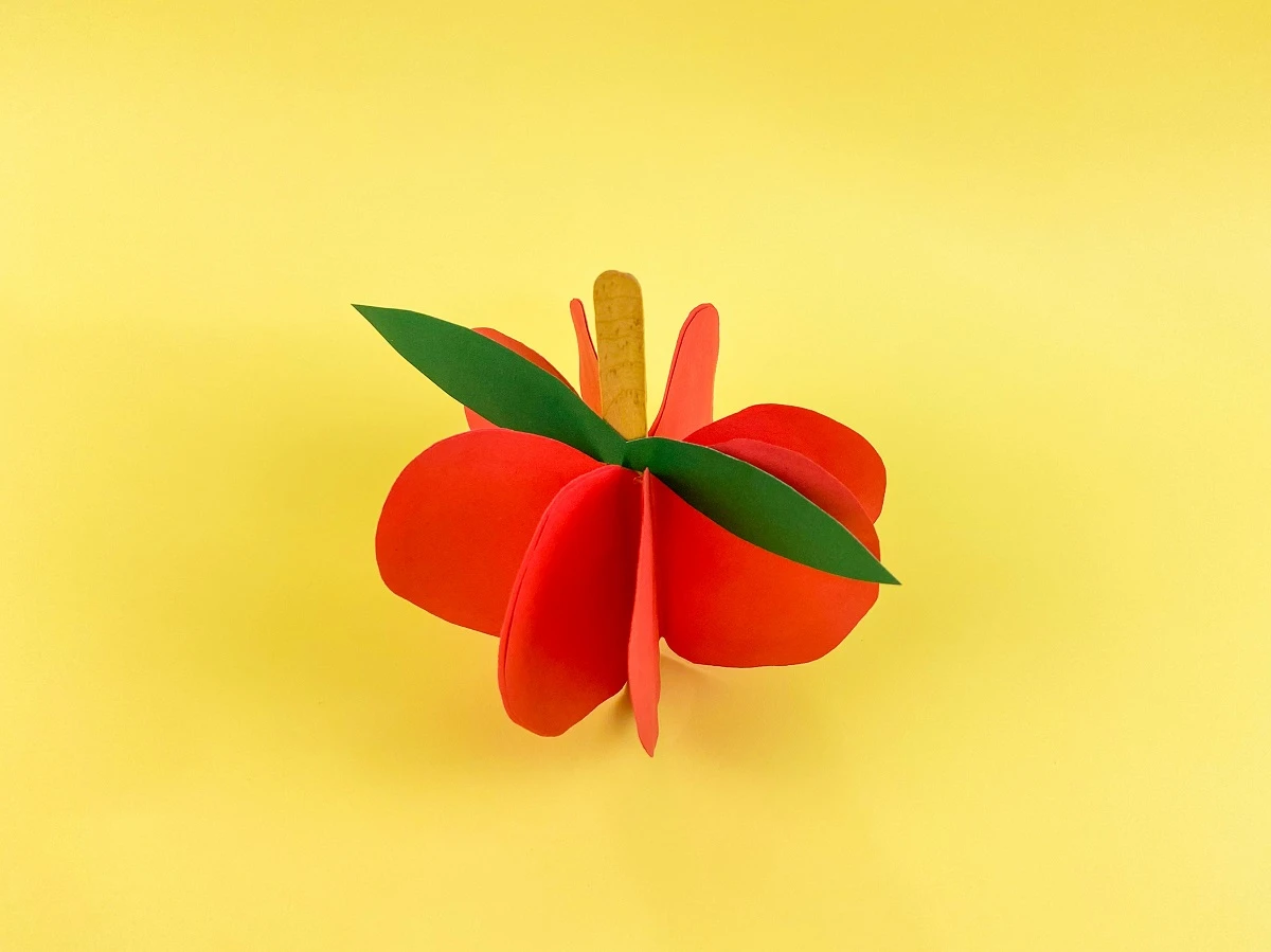 Angled view from the top showing the completely assembled apple made with folded pieces of red paper glued together around a popsicle craft stick. A green paper apple leaf is glued to the craft stick. The craft is sitting on a yellow background.