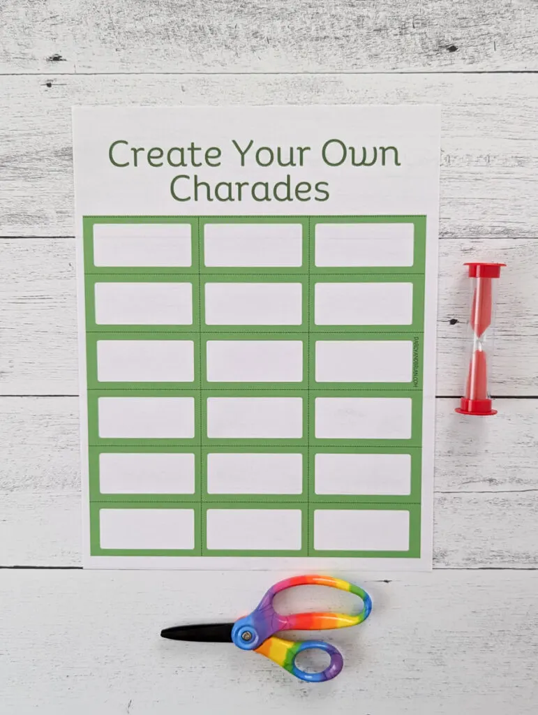 A printed out page of blank charades cards to create your own. A pair of rainbow handled scissors and a red sand timer lay next to the paper.
