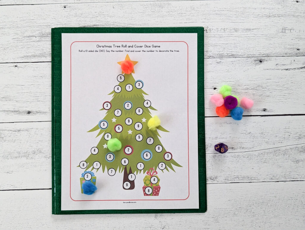 Christmas tree roll and cover game page for numbers 0-9 printed out. A few pom poms cover numbers on the tree and presents. A pile of different colored pom poms lays next to the paper and a 10 sided die.