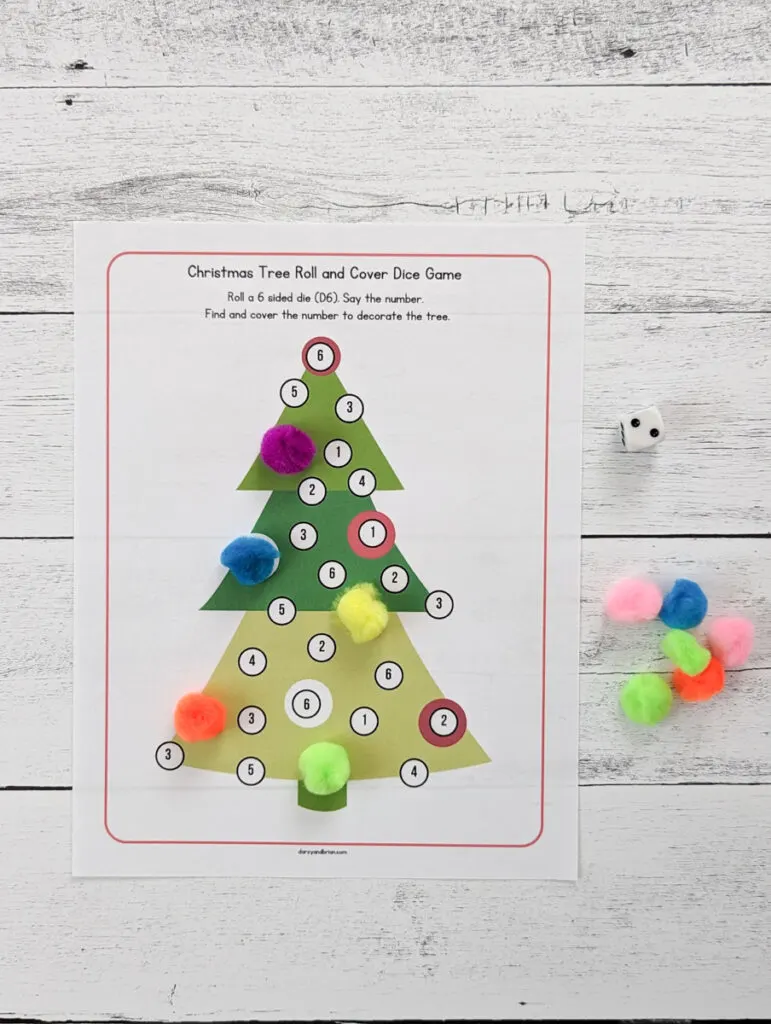 Overhead view of Christmas tree dice game printed out with various colorful pom poms covering numbers that have been rolled. Tree has numbers one through six on it. A six sided die with two pips is facing up near a pile of pom poms.