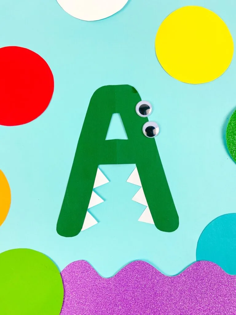 Completely finished alphabet alligator craft. Bright colored shapes are arranged around it for decoration.