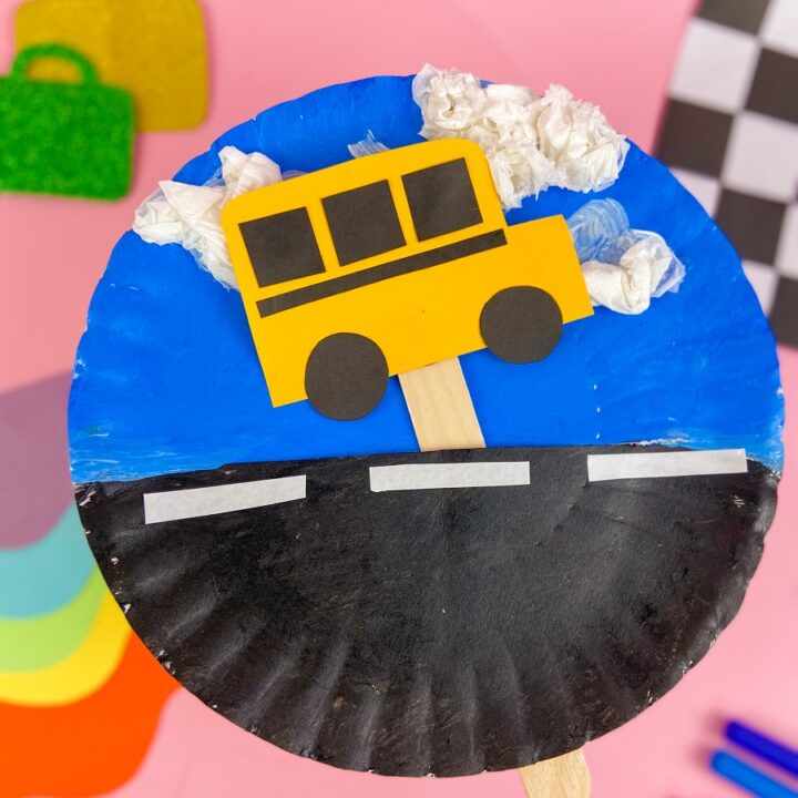 Moving school bus paper plate craft project completely assembled. Background of photo is styled to show the finished project - pink background, different colors of paper, a checkered flag.