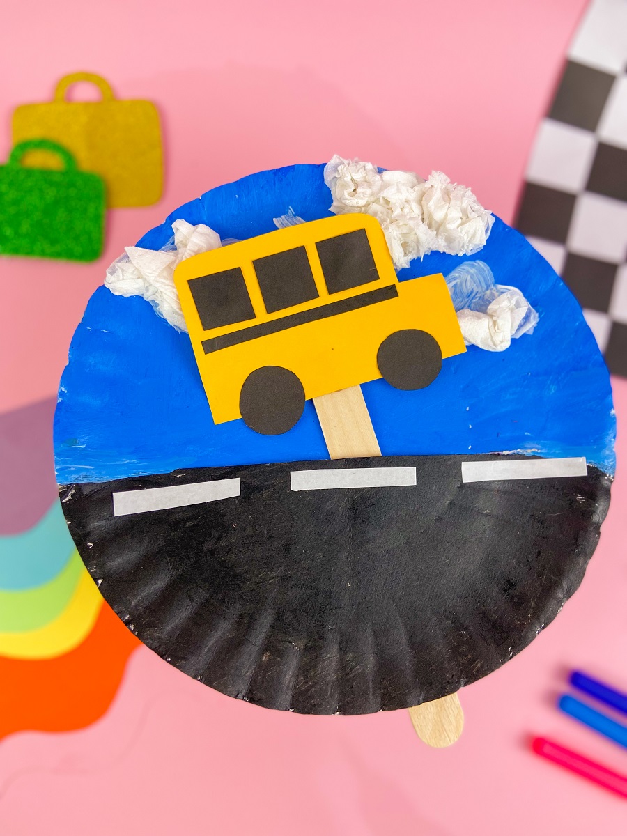 Moving school bus paper plate craft project completely assembled. Background of photo is styled to show the finished project - pink background, different colors of paper, a checkered flag.