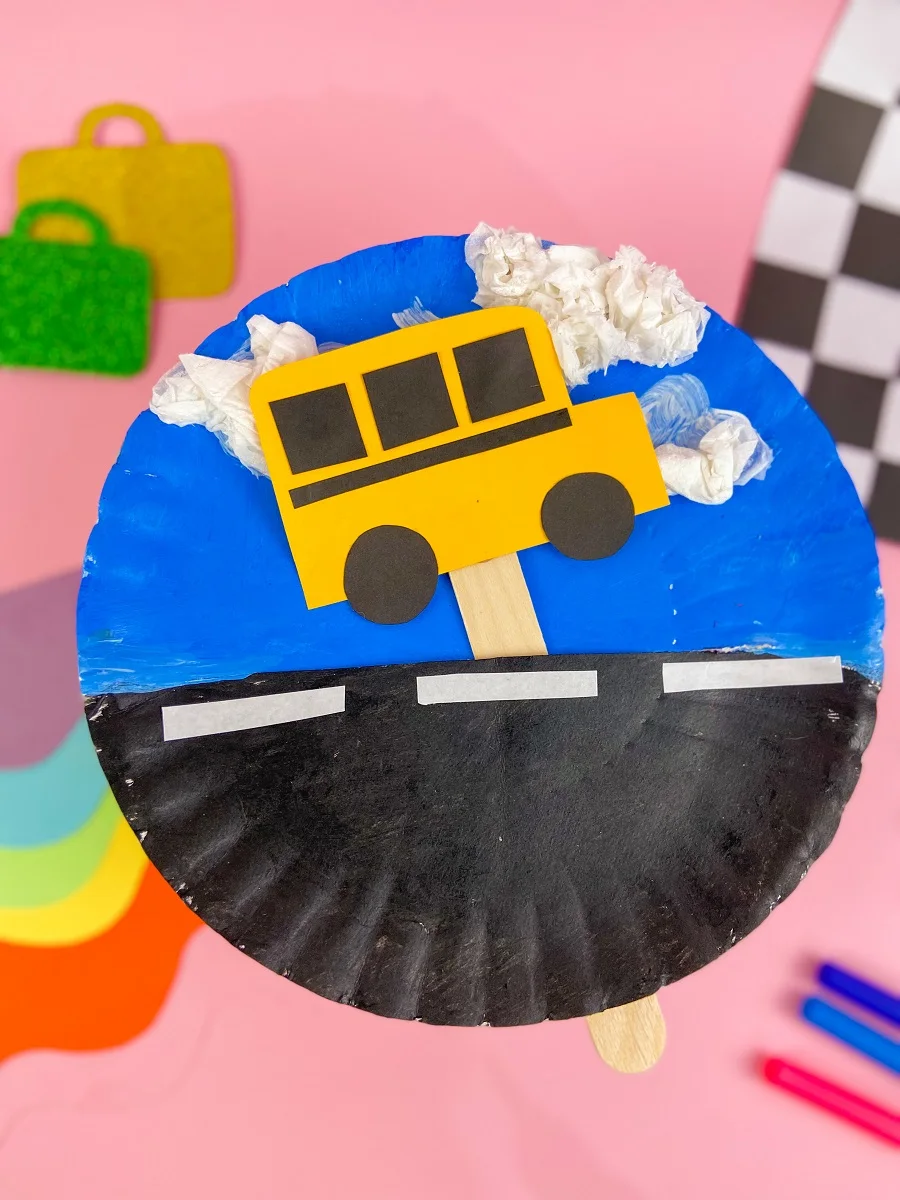 Moving school bus paper plate craft project completely assembled. Background of photo is styled to show the finished project - pink background, different colors of paper, a checkered flag.