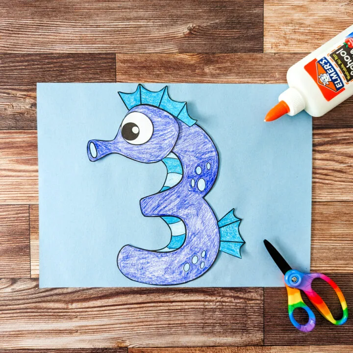 Seahorse template pieces assembled and arranged on light blue construction paper to look like a number 3. Bottle of glue and kid scissors lay nearby on the right.