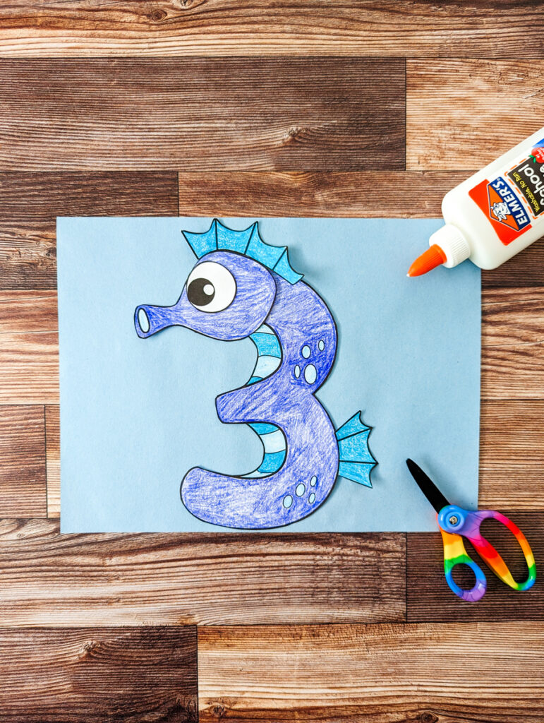 Seahorse template pieces assembled and arranged on light blue construction paper to look like a number 3. Bottle of glue and kid scissors lay nearby on the right.