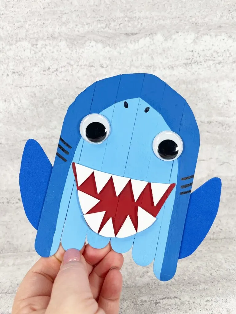 White woman's hand holding up shark made out of blue painted popsicle sticks and craft foam. It also has a red painted mouth open with pointy white craft foam teeth and googly eyes that make it cute.