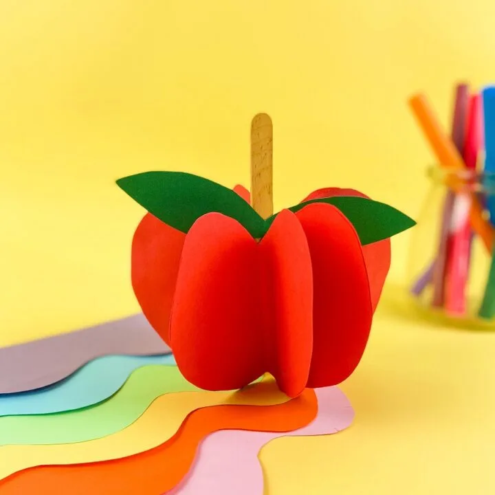 3D apple standing up on desk covered with assorted colors of paper and markers on a yellow background. Apple made with red paper, popsicle stick, and green paper.