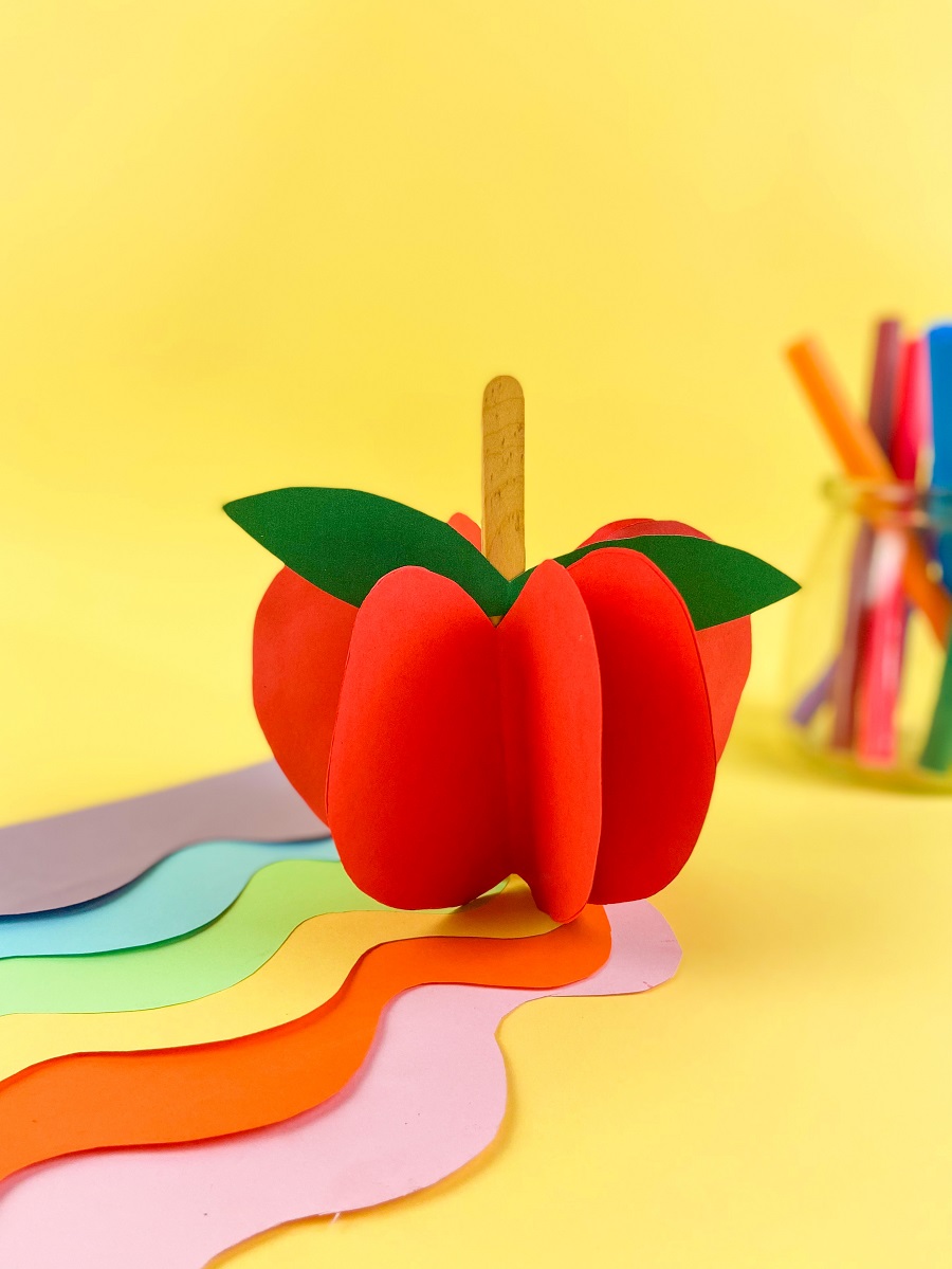 3D apple standing up on desk covered with assorted colors of paper and markers on a yellow background. Apple made with red paper, popsicle stick, and green paper.