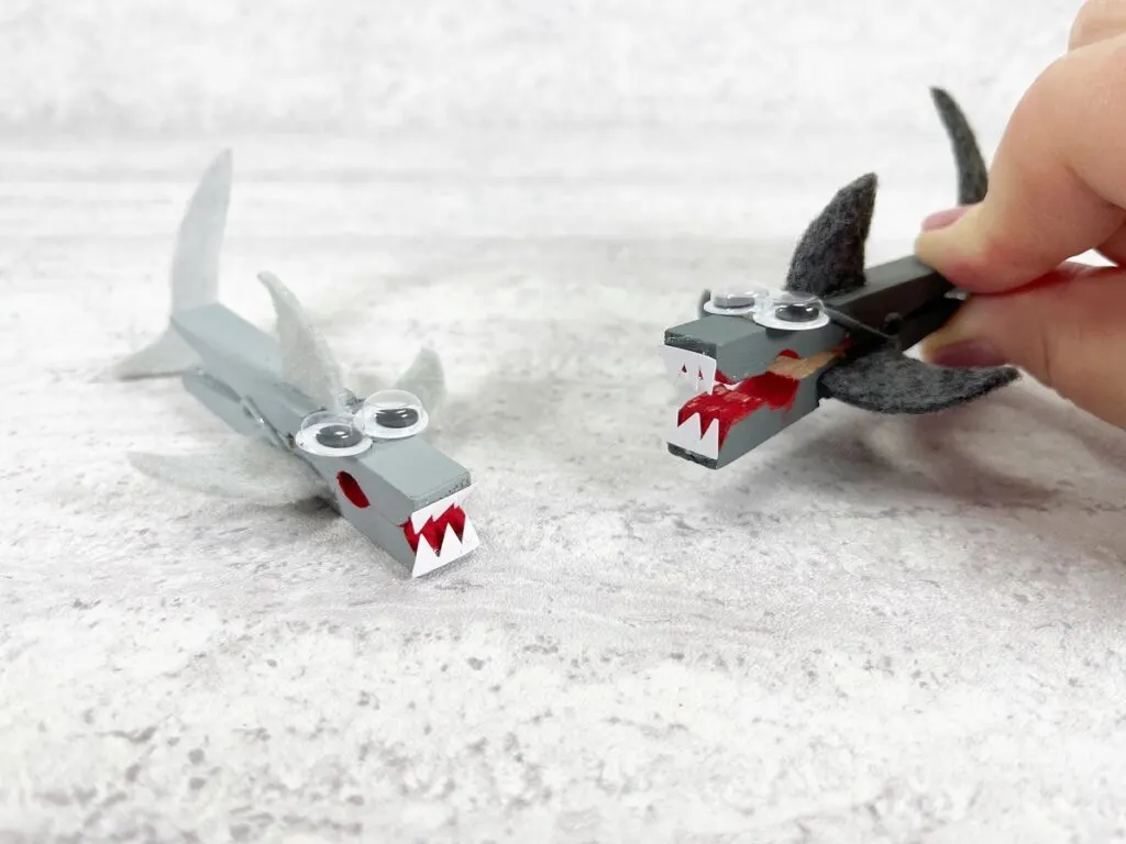 Two completed clothespin shark crafts. Left one is painted light gray. Right one is painted dark gray and is being held with its mouth open. 