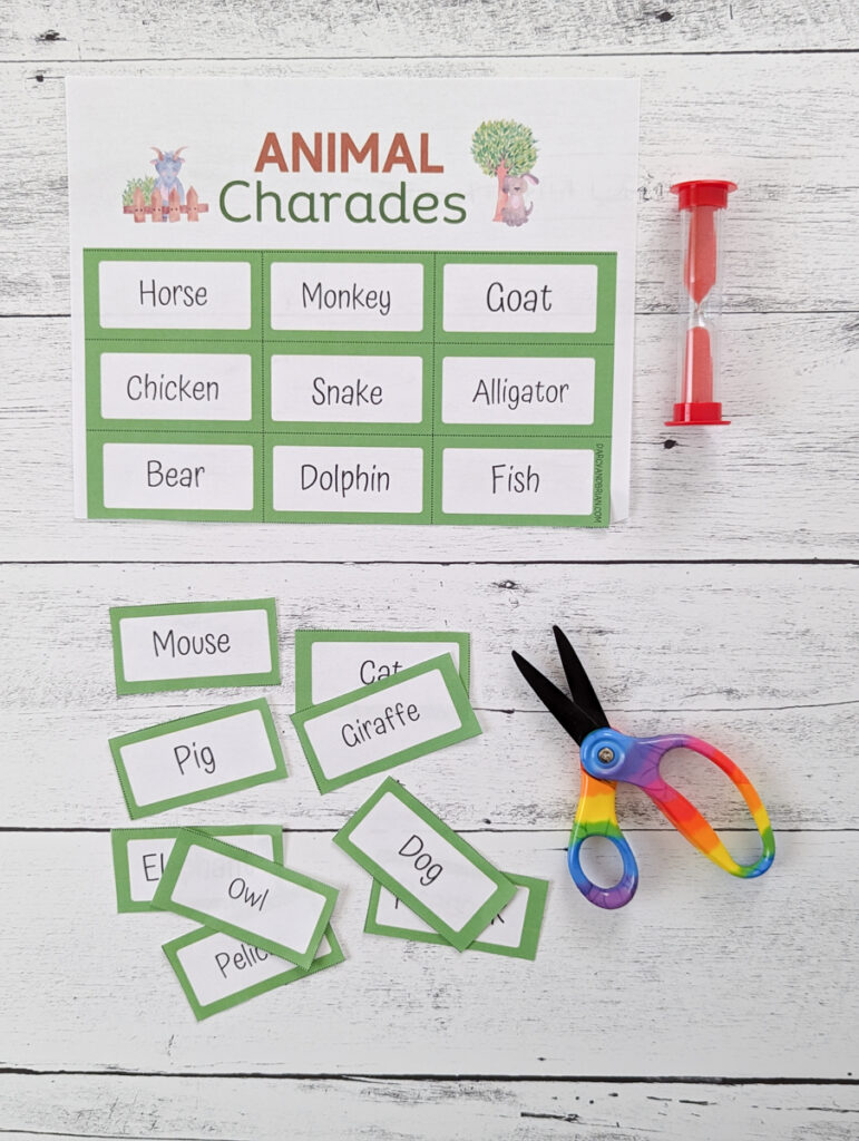 Half a sheet of animal charades is near the top next to a red sand timer. The rest of the word cards are cut apart and laying scattered near a pair of scissors.