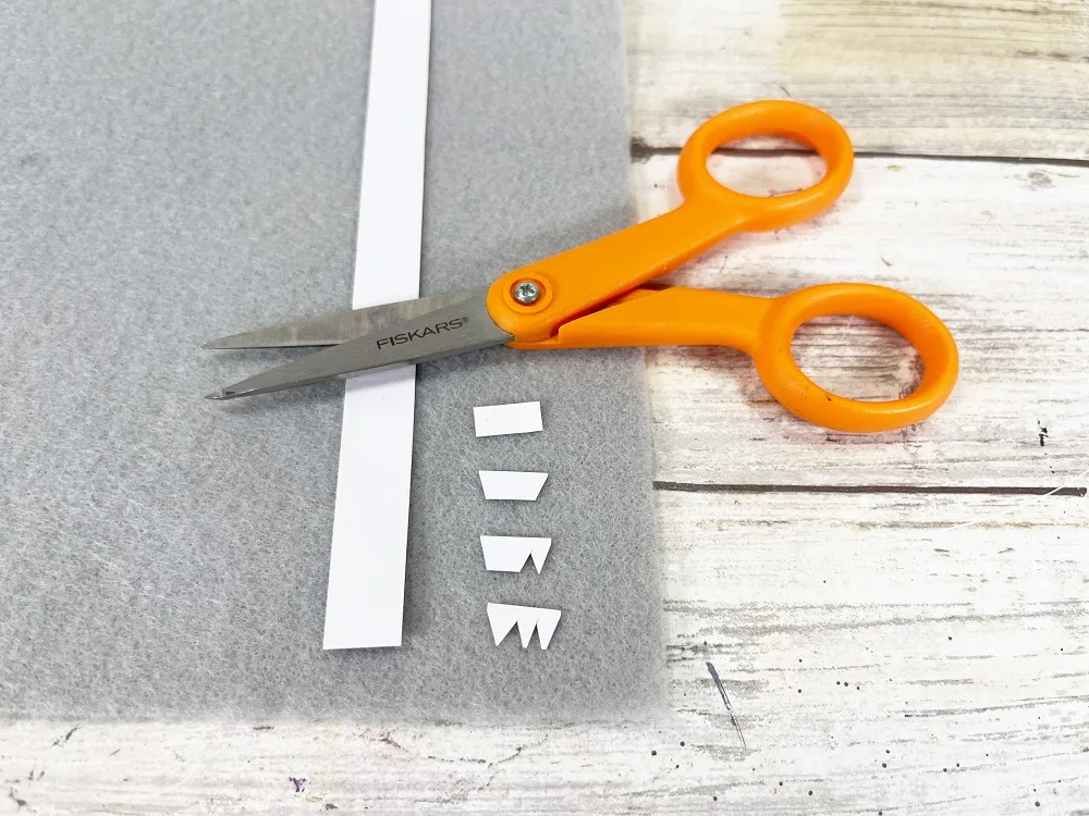 Strip of white paper laying on top of gray craft felt. Some small rectangular pieces have been cut off of the strip. Some have little triangles cut out. Small pair of orange handled scissors laying across the paper.