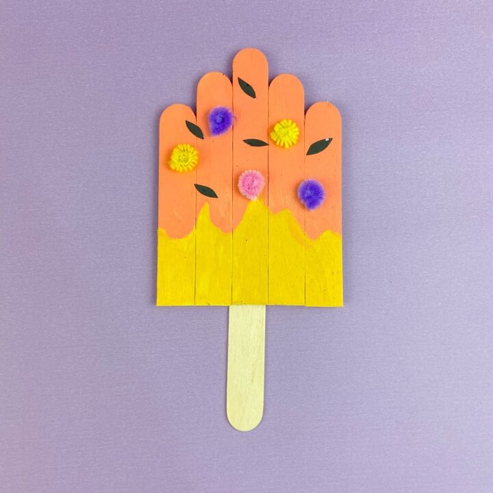 Painted craft sticks glued together to create a popsicle craft. Popsicle is decorated with pieces of paper and pipe cleaners. Completed projects lays on a purple background.
