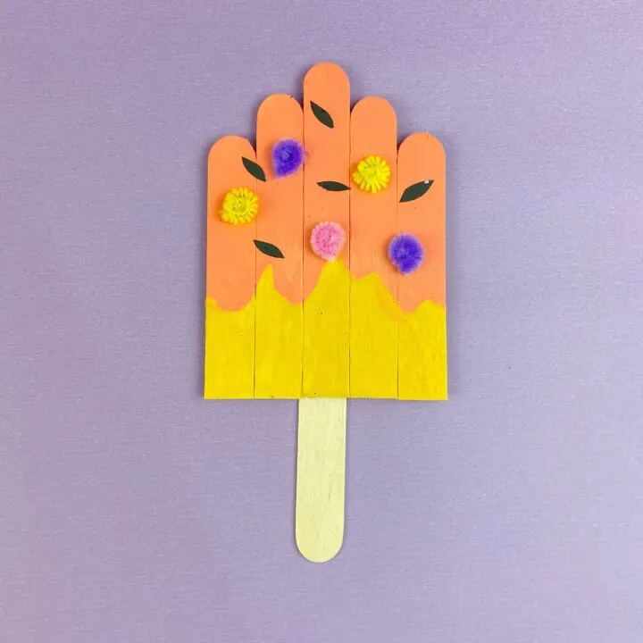 Painted craft sticks glued together to create a popsicle craft. Popsicle is decorated with pieces of paper and pipe cleaners. Completed projects lays on a purple background.